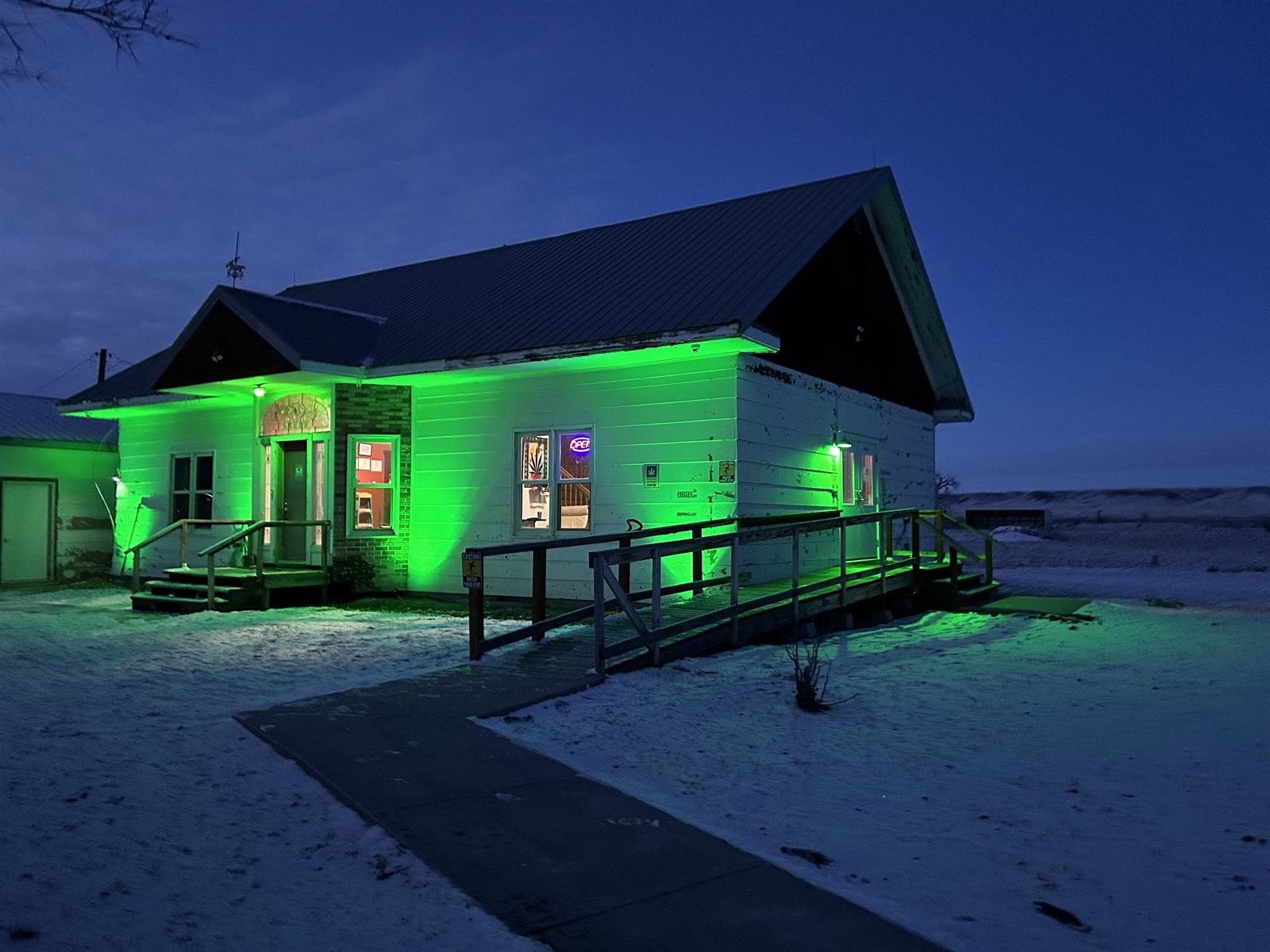 Highline Apothecary at night, the white building washed in green light, with snow on the ground and the ramp leading up to the door